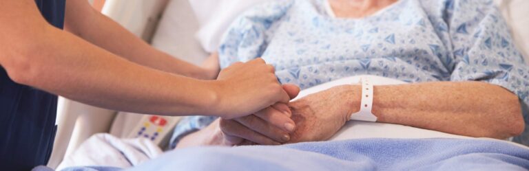 A person holds the hand of an elderly patient lying in a hospital bed wearing a blue gown and a wristband.