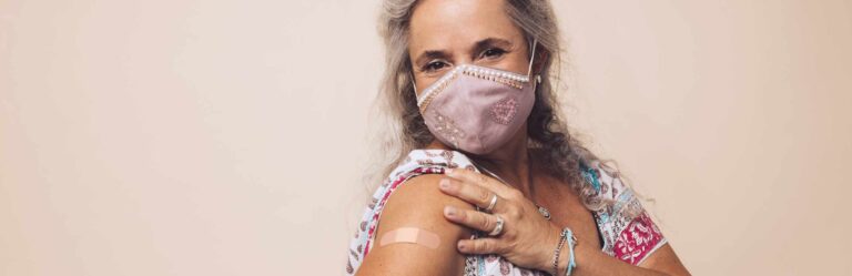 An older adult with gray hair wearing a face mask shows an adhesive bandage on their upper arm, indicating they have received a vaccination.