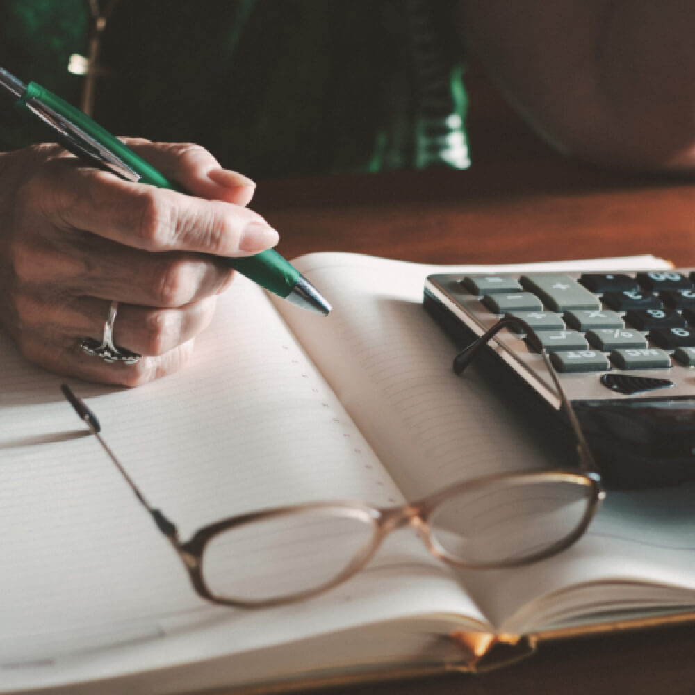 A pair of glasses sits on a notepad next to a calculator while someone writes a note.