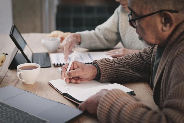 Two people sit at a table working on laptops and writing in notebooks, with a coffee cup placed nearby.