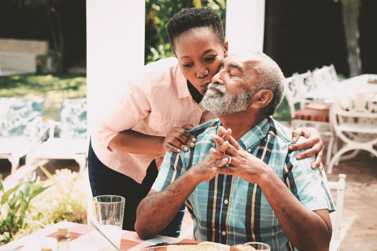 A woman kisses the cheek of a man seated at an outdoor dining table. They both appear happy. There are glasses and a plate on the table and patio furniture in the background.