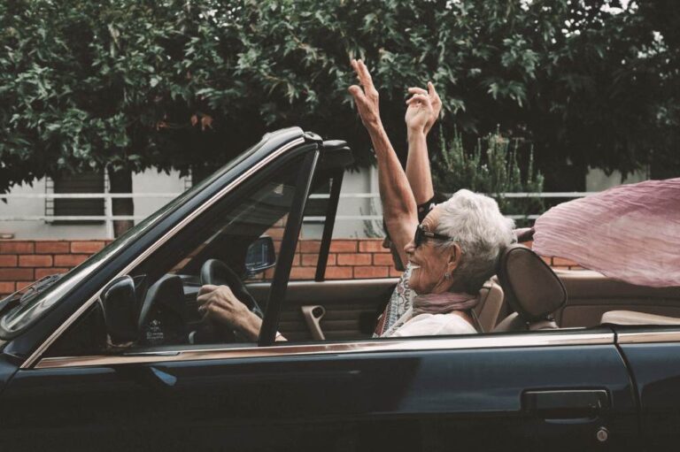 Senior woman with short gray hair lifts her hand in joy as she drives a convertible car with the top down.